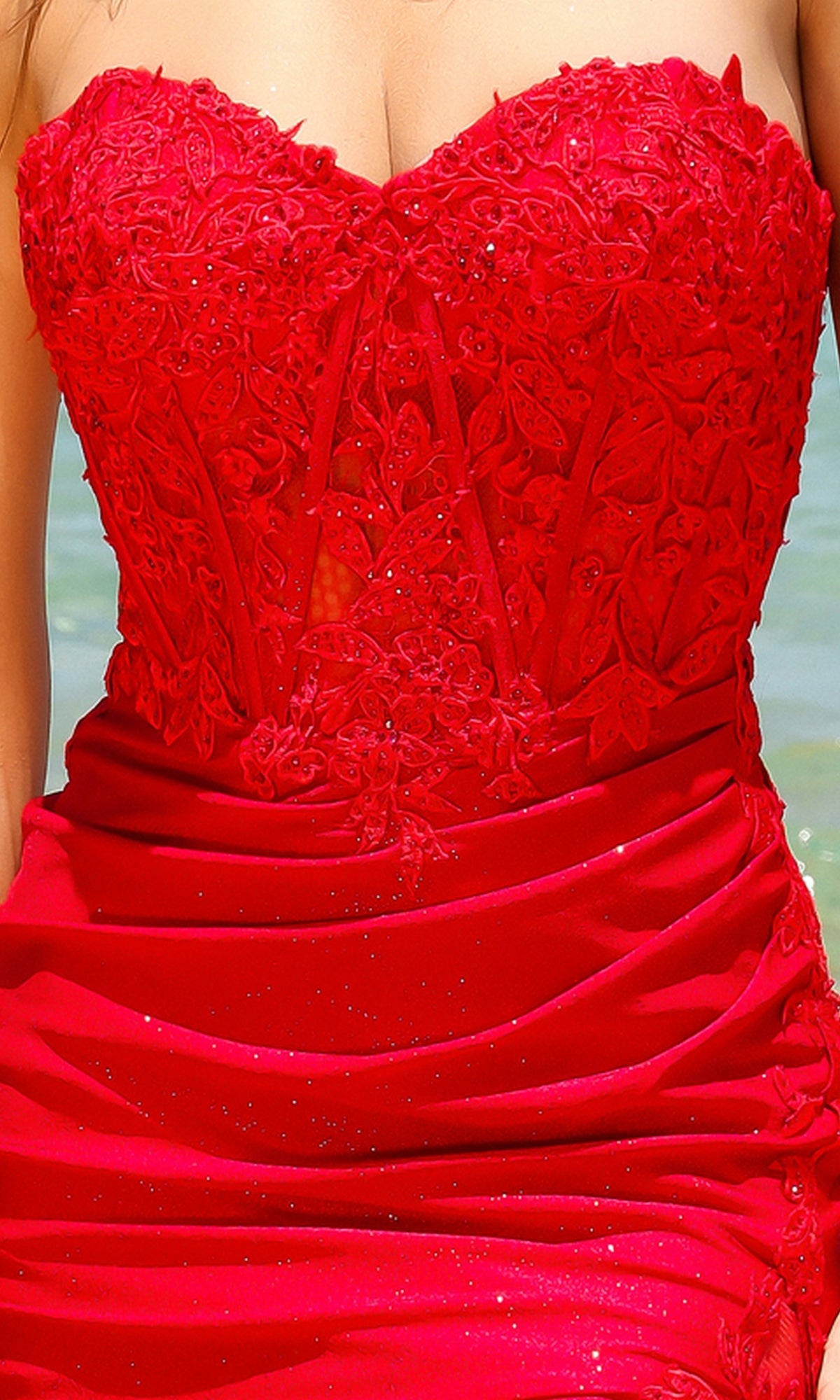 A person wearing the Amelia Couture 7031 Strapless Long Prom Dress, featuring floral lace and sparkling accents, stands outdoors near water.